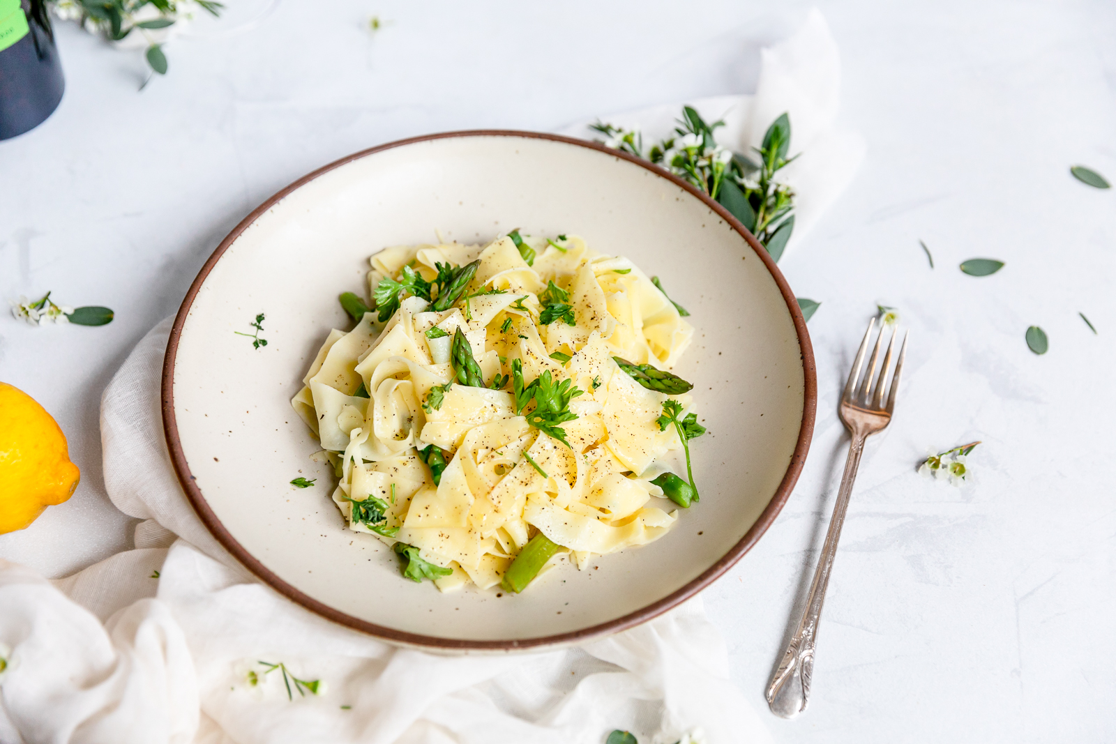 Plate of wide pappardelle pasta with asparagus, garnished with fresh herbs and cracked black pepper, served on a white plate with a fork on the side.