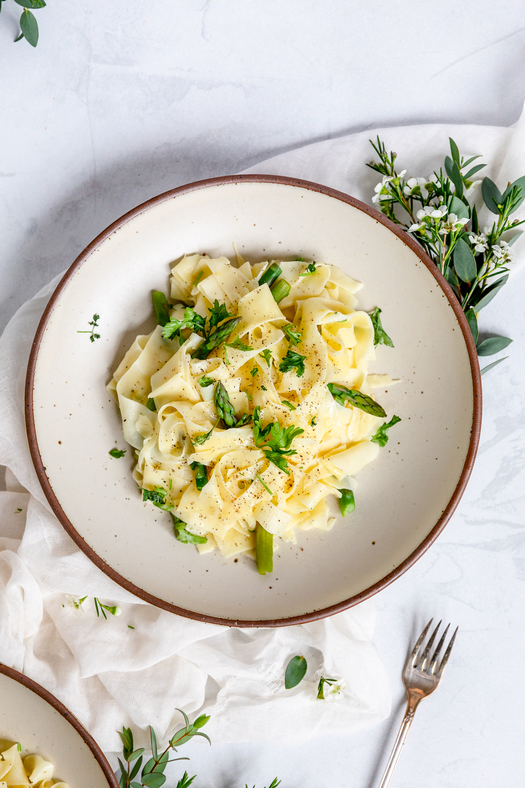 Overhead view of a plate of pappardelle pasta with asparagus, topped with fresh herbs and grated cheese, served with a fork on the side.