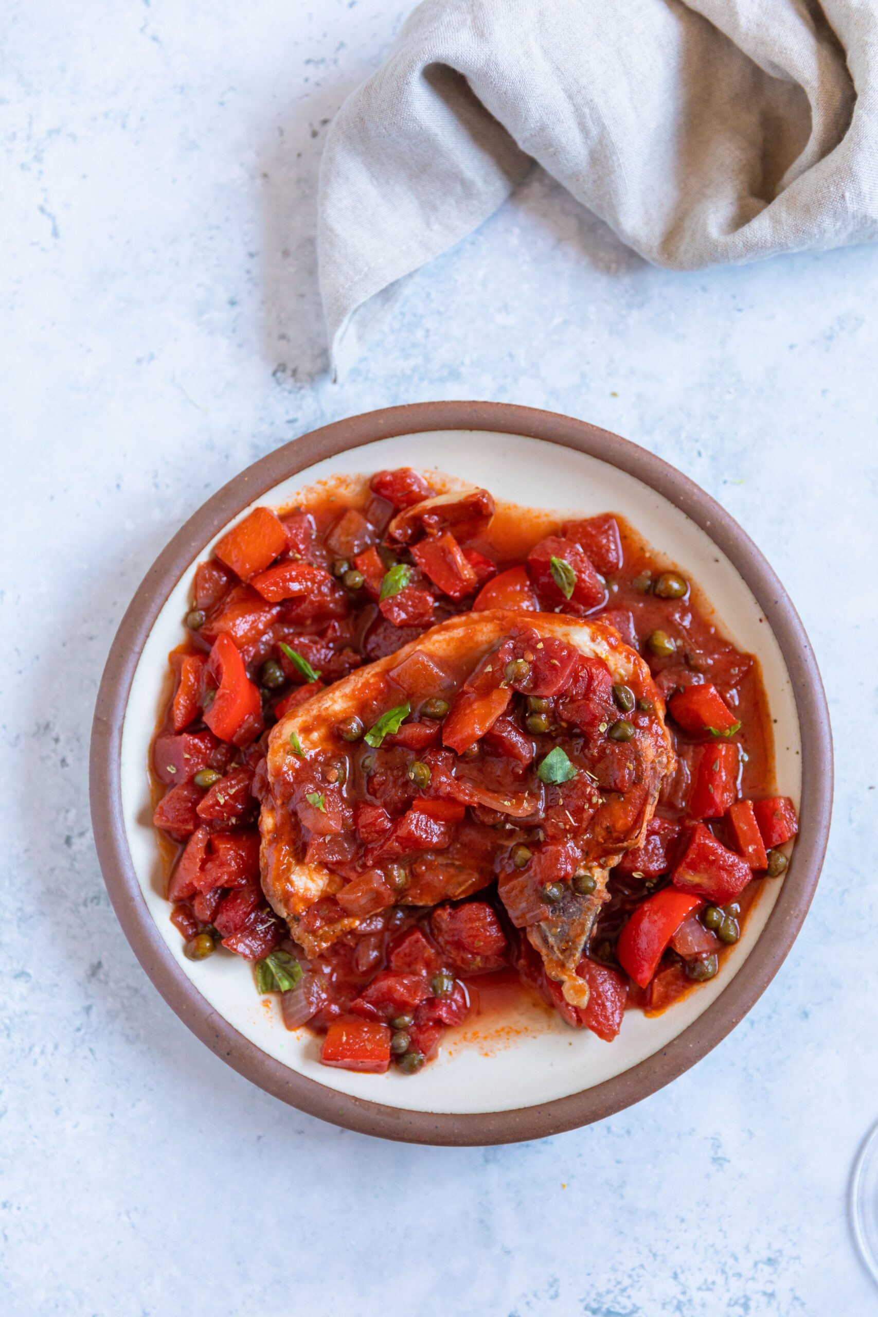 Overhead of a tomato-based puttanesca sauce over a pork chop on a white plate and a light tan colored fabric napkin off to the side.