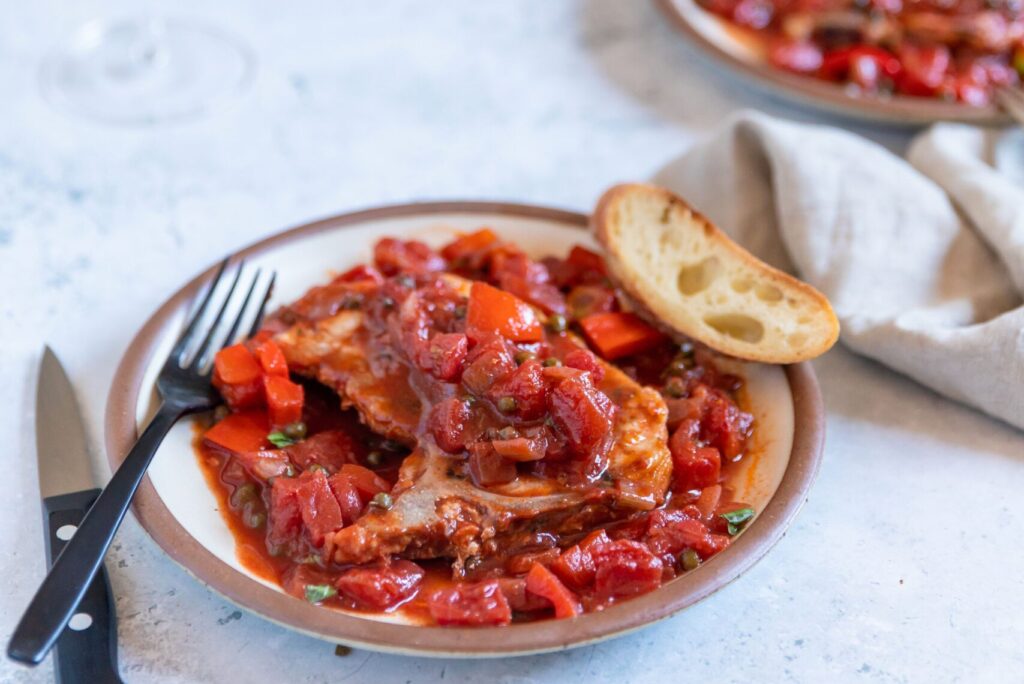 Tomato-based puttanesca sauce over a pork chop on a white plate with a piece of baguette on the side and a black fork and knife. Two glasses of red wine and a case of small white flowers in the background.