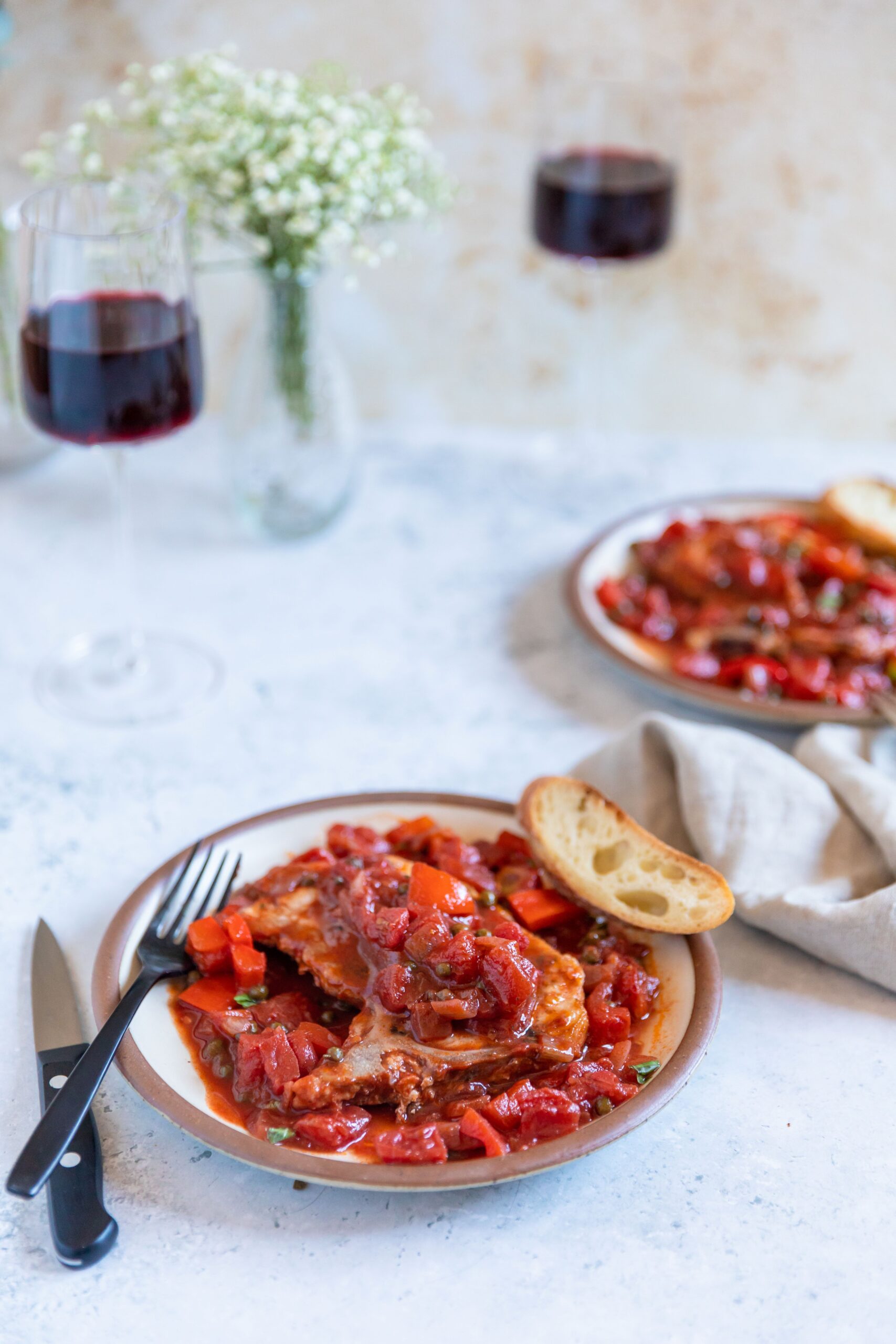Tomato-based puttanesca sauce over a pork chop on a white plate with a piece of baguette on the side and a black fork and knife. Two glasses of red wine and a case of small white flowers in the background.