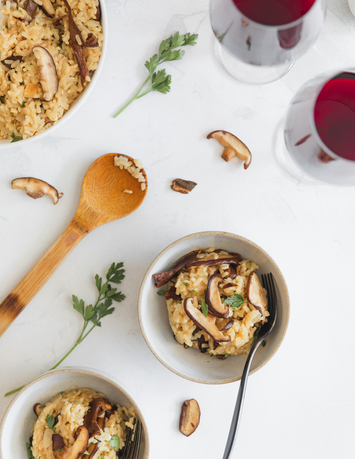 Two bowls of creamy mushroom risotto topped with sautéed mushroom slices and parsley, set on a pale surface. A larger serving bowl of risotto, a wooden serving spoon and two glasses of red wine are nearby.