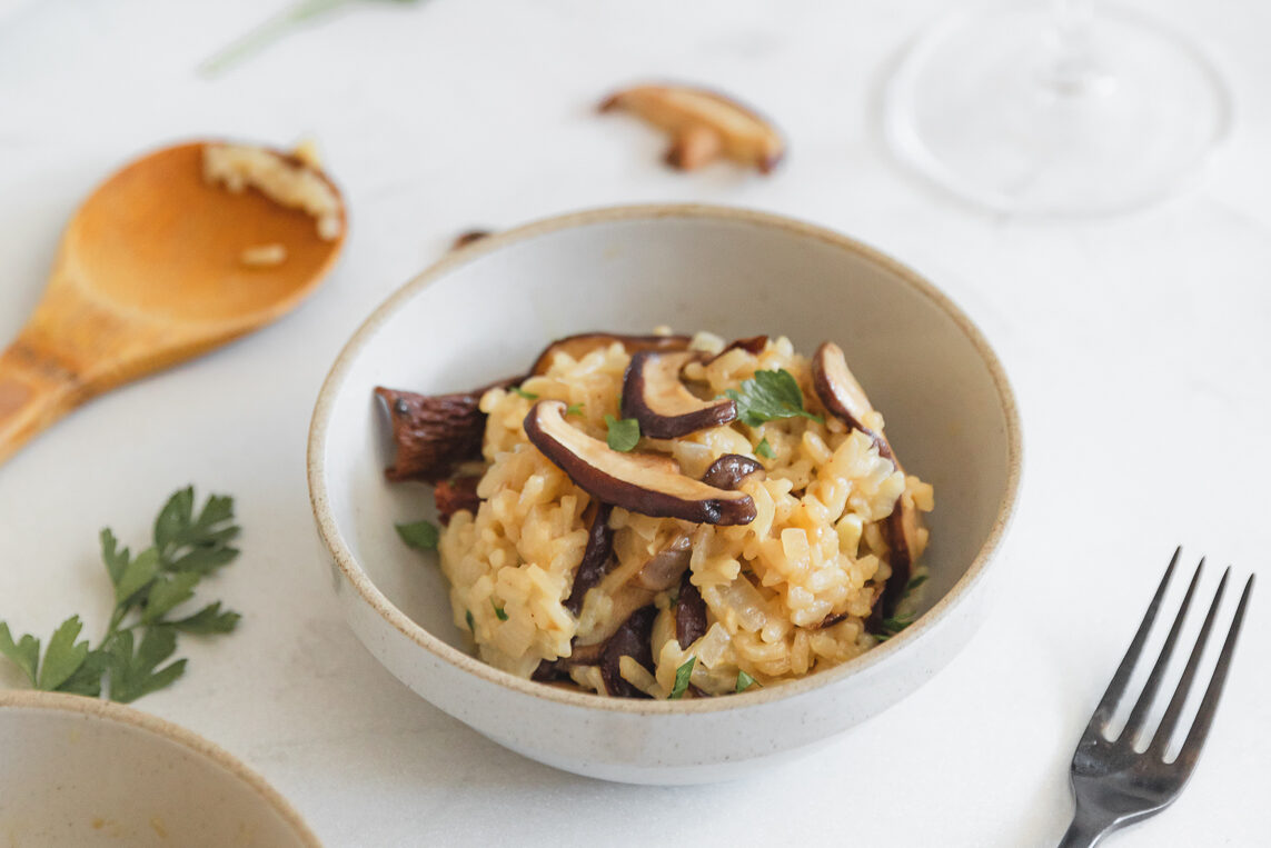 Bowl of creamy mushroom risotto topped with sautéed mushrooms and parsley, with a wooden serving spoon and fork on the side.