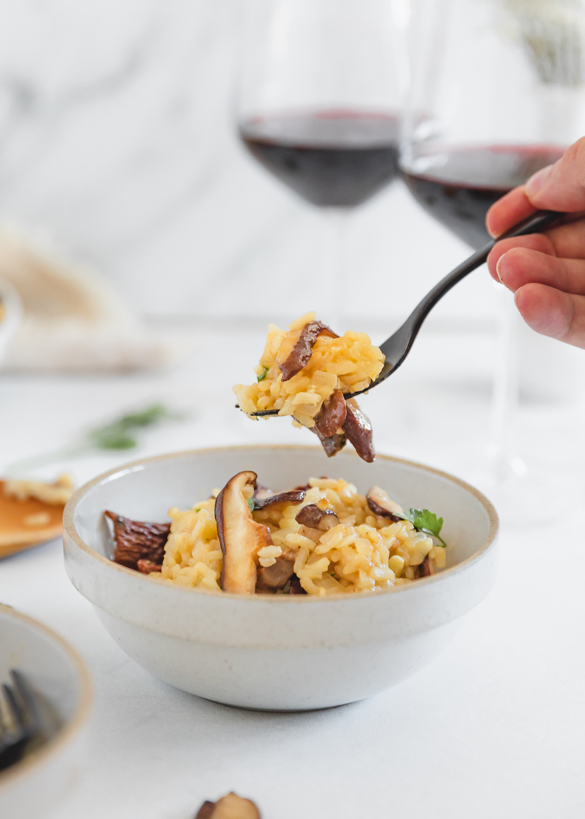 A bowl of creamy risotto topped with mushrooms and parsley, with a hand holding a black fork that had a bite of the risotto on it. Two glasses of red wine are nearby in the background.