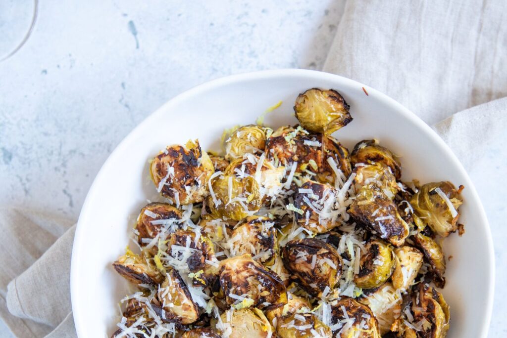 Overhead of a partial white bowl of halved and roasted brussels sprouts topped with grated parmesan cheese on a light background.