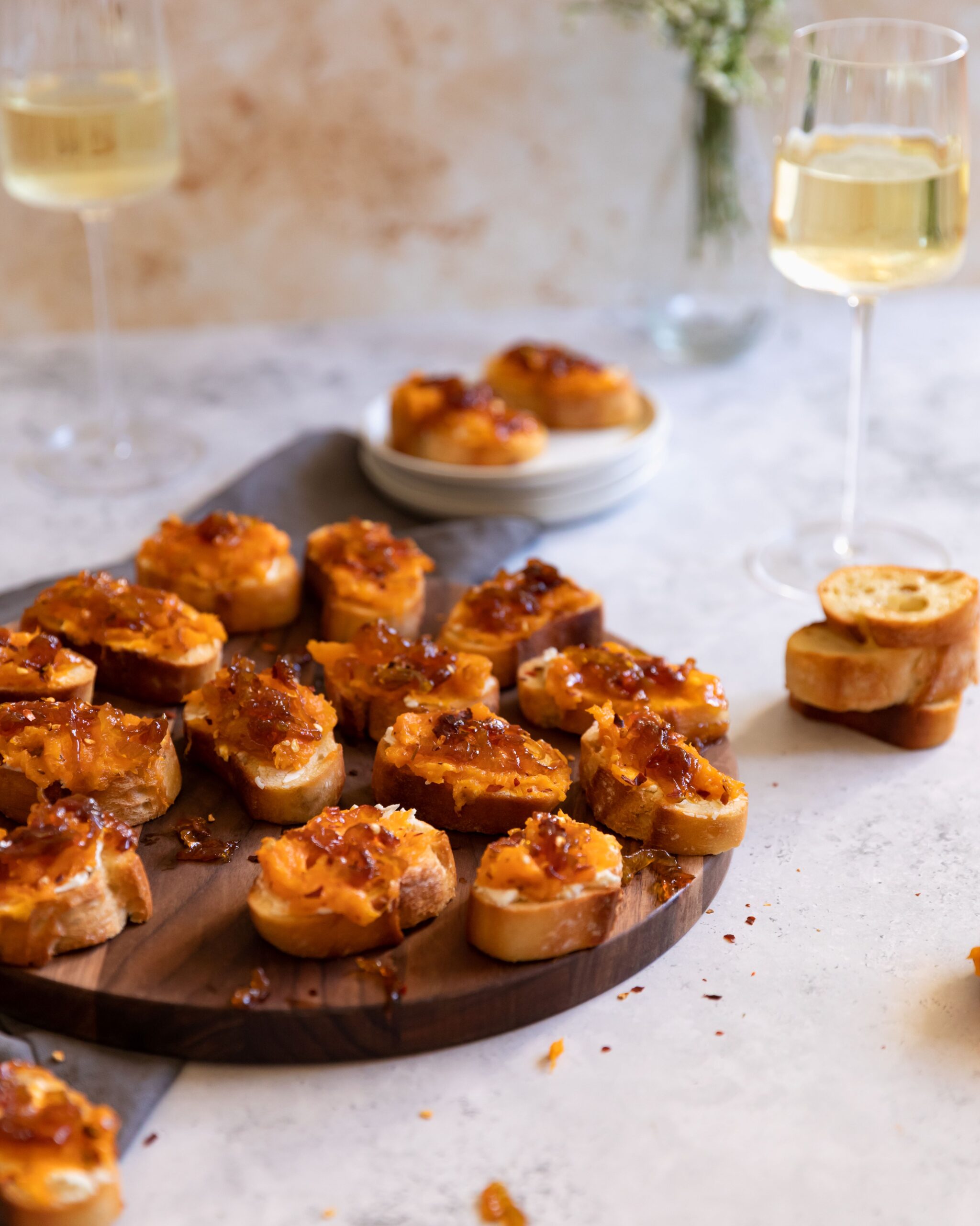 Many toasted baguette slices each topped with spreadable ricotta cheese, mashed butternut squash, and caramelized onions placed on a round, dark wood cutting board sitting on a light counter surface. Two additional topped slices are on a small white plate behind the cutting board and a few additional slices of toasted, but not topped, bread sit off to the side. Two glasses of white wine and a clear glass vase of greenery and white flowers are all in the background.