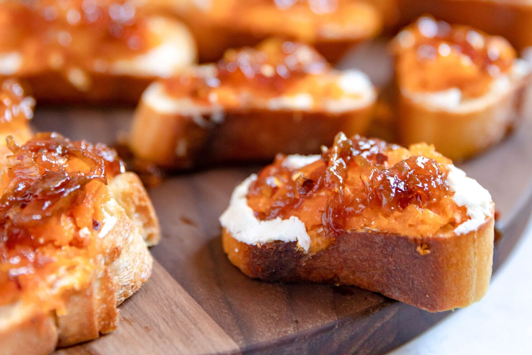 Close up of multiple toasted baguette slices each topped with spreadable ricotta cheese, mashed butternut squash, and caramelized onions placed on a dark wood cutting board.