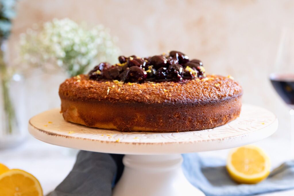 Close-up of a golden-brown cake topped with dark cherry compote, displayed on a white cake stand with a blurred background.