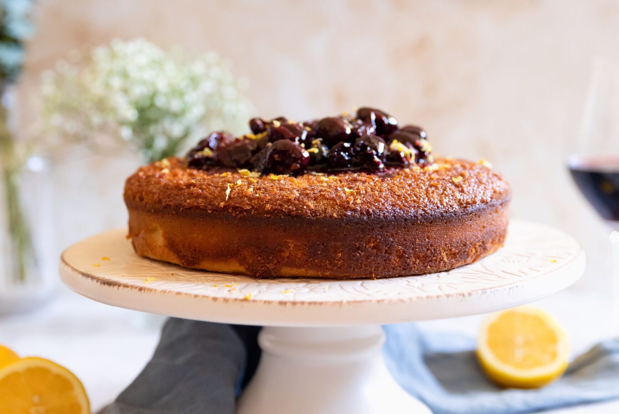 Close-up of a golden-brown cake topped with dark cherry compote, displayed on a white cake stand with a blurred background.