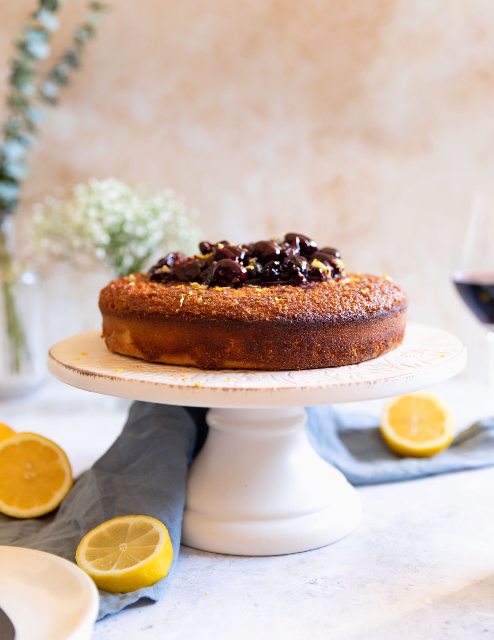 Side view of a golden-brown cake topped with dark cherry compote, displayed on a white cake stand with lemon halves, a glass of red wine, and flowers on a light background.