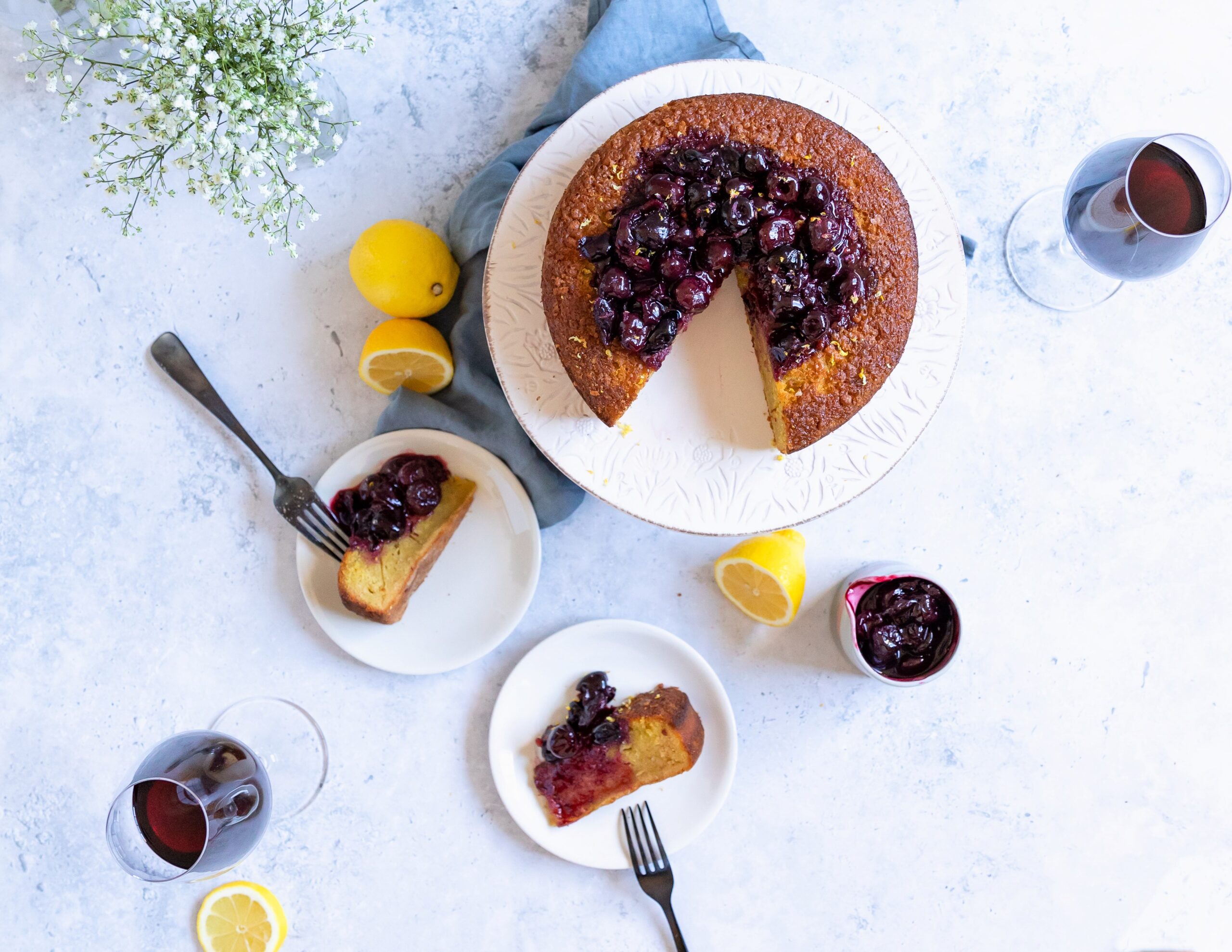 Overhead view of a round cake topped with cherry compote on a white stand, with slices served on plates alongside forks, lemon halves, glasses of red wine, and flowers on a light background.