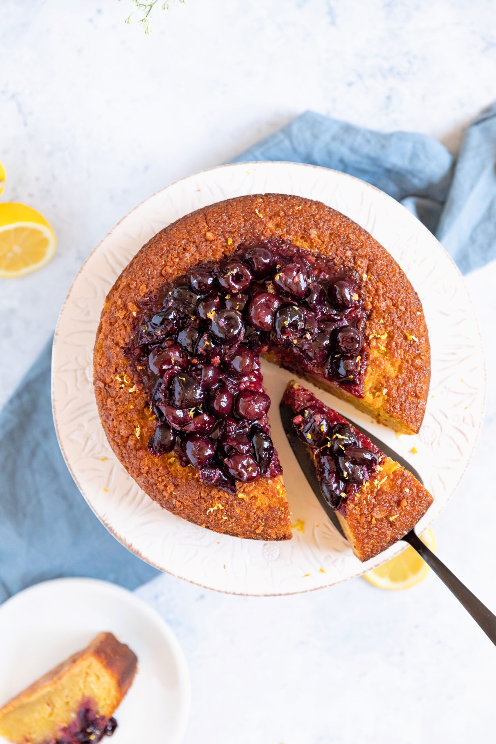 Top view of a round cake topped with cherry compote, with a slice being lifted from the cake on a server, set on a white cake stand with lemon slices and a blue cloth nearby.