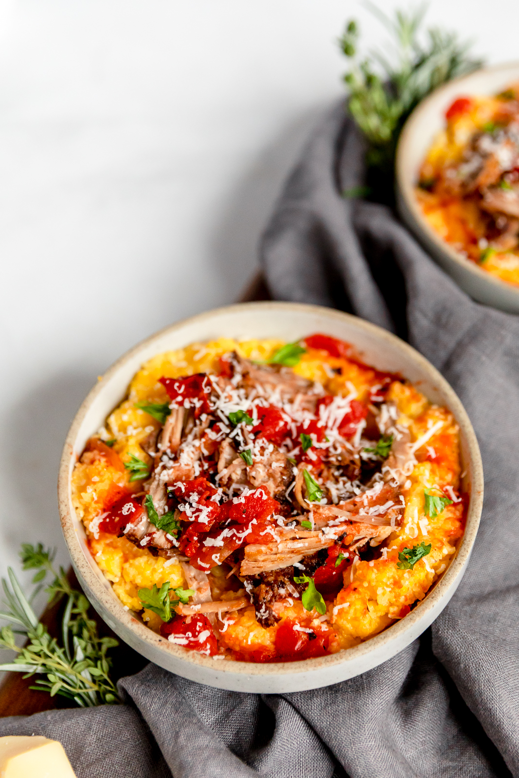 Overhead one full bowl, and another off to the side, of pulled pork with tomatoes over polenta, garnished with fresh green herbs and grated parmesan cheese, with a light gray cloth napkin and additional green herbs in the background.