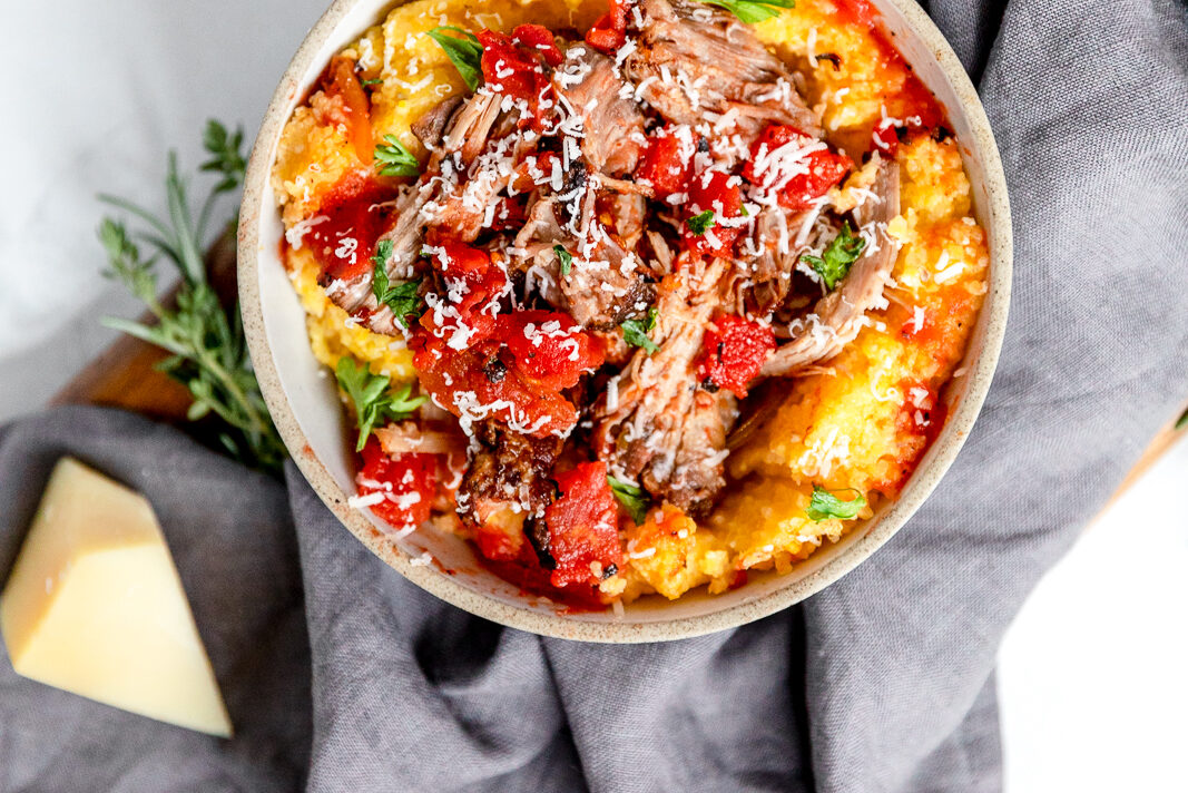 Overhead of a bowl of pulled pork with tomatoes over polenta, garnished with fresh green herbs and grated parmesan cheese, with a light gray cloth napkin in the background.