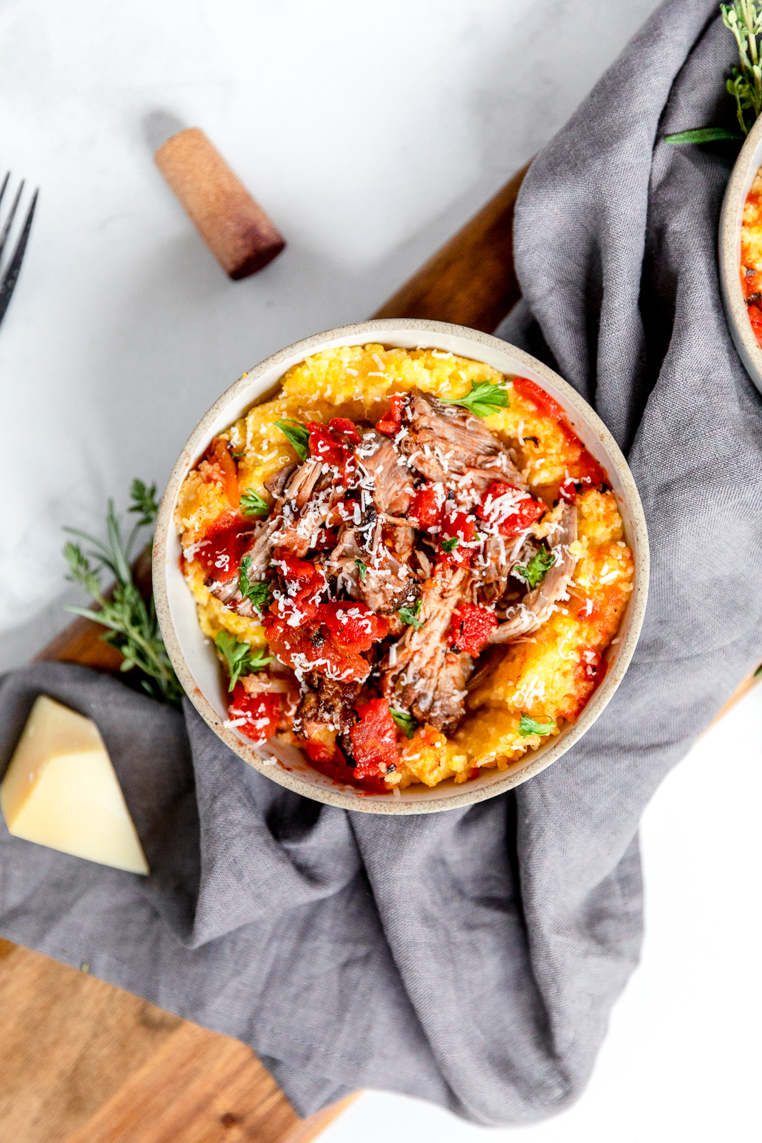 Overhead of a bowl of pulled pork with tomatoes over polenta, garnished with fresh green herbs and grated parmesan cheese, with a light gray cloth napkin, a wood cutting board, and a red wine cork in the background.