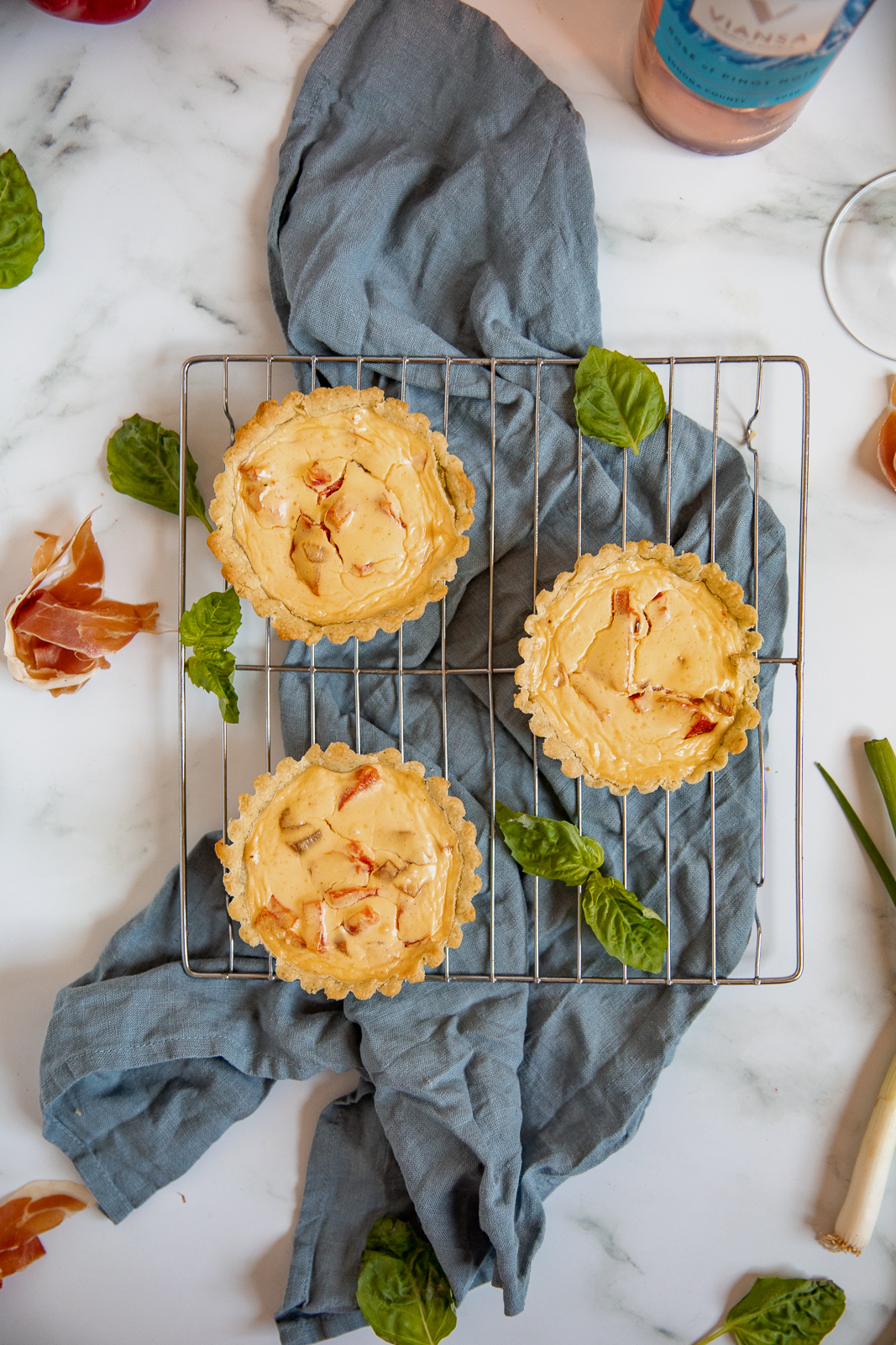 Overheard of three mini tarts with red peppers and goat cheese sitting on a metal cooling rack on top of a gray blue linen napkin. Basil leaves, a whole green onion, prosciutto, and the bottom of a bottle of rosé wine are also visible in the surrounding space.