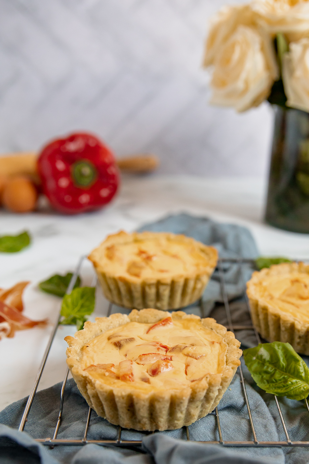 Close up of three mini tarts with red peppers and goat cheese sitting on a metal cooling rack on top of a gray blue linen napkin. Basil leaves surround the tarts, while a red pepper, egg, rolling pin, and vase of cream roses are visible in the background.