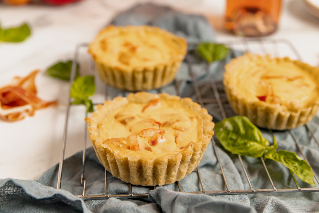 Close up of three mini tarts with red peppers and goat cheese sitting on a metal cooling rack on top of a gray blue linen napkin. Basil leaves and the bottom of a bottle of rosé wine are also visible in the surrounding space.