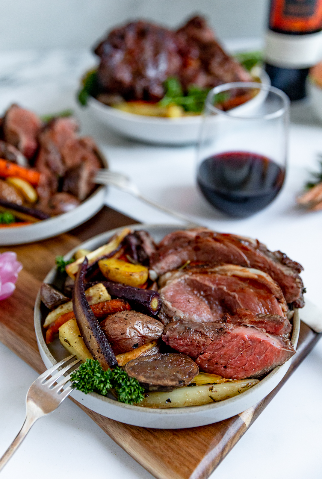 Three dishes of lamb cooked rare over a bed of colorful vegetables in a white shallow bowl with a stemless glass of red wine and the wine bottle in the background.