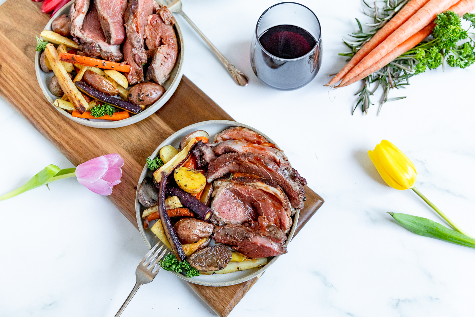 Overhead shot of two dishes of lamb cooked rare over a bed of colorful vegetables in a white shallow bowl with a stemless glass of red wine in the background.