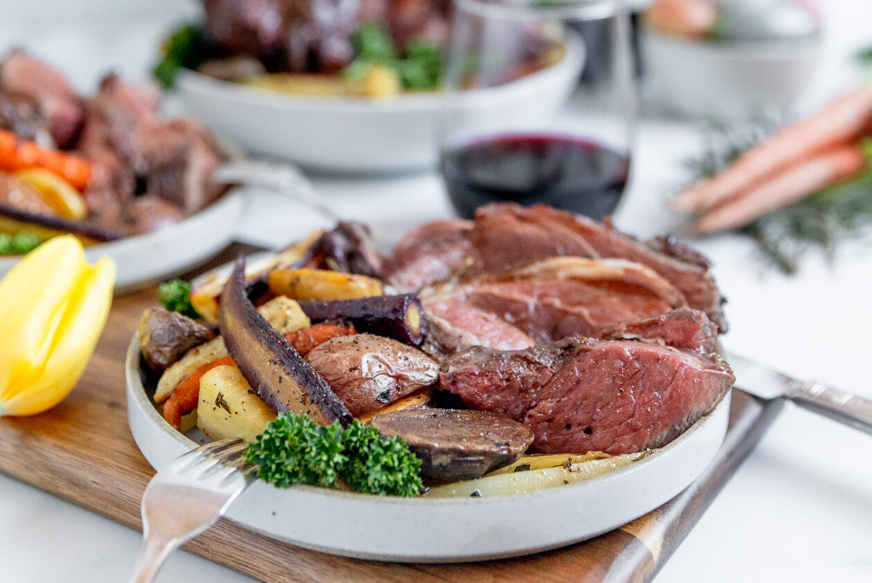 Lamb cooked rare over a bed of colorful vegetables in a white shallow bowl with a stemless glass of red wine in the background.