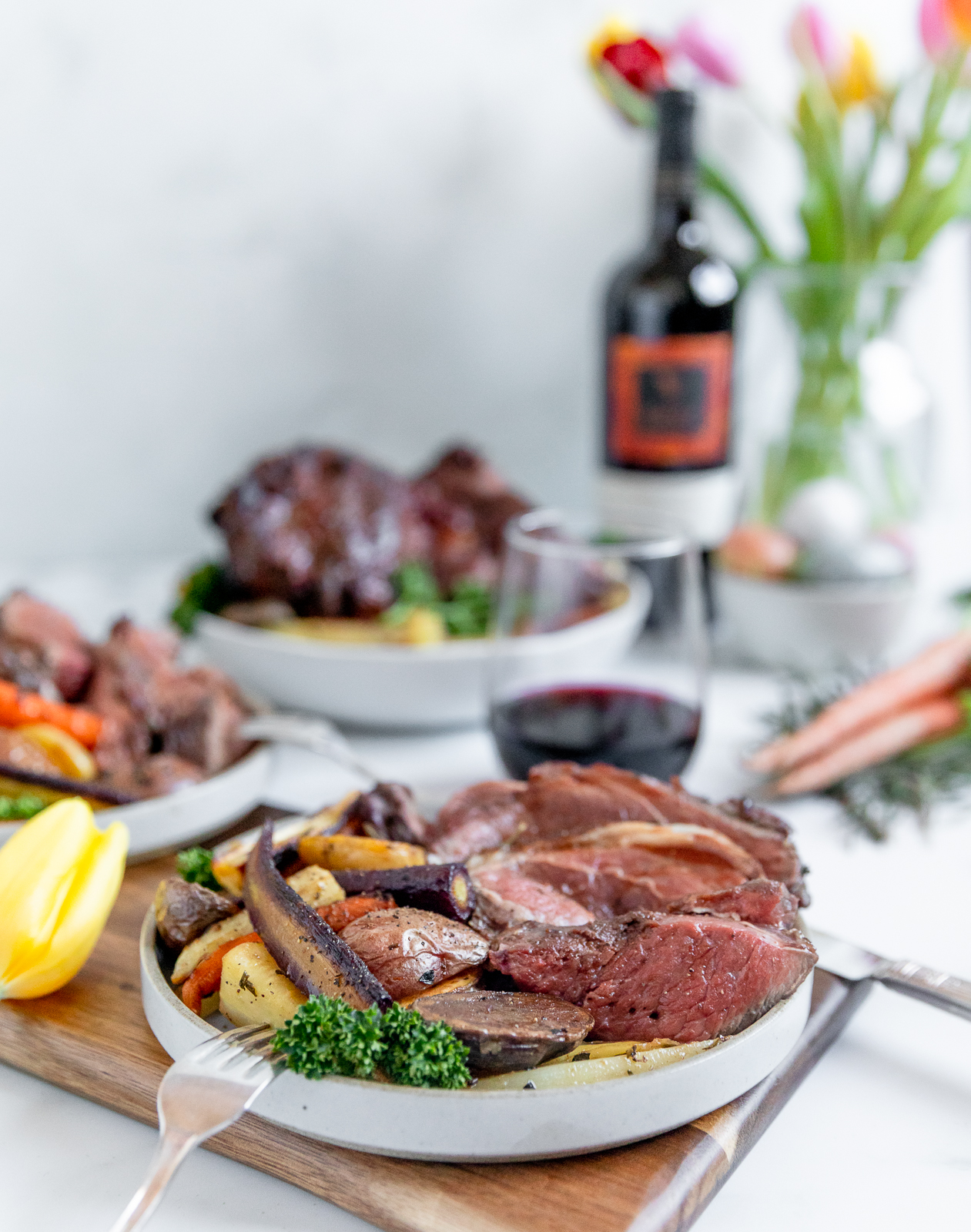 Lamb cooked rare over a bed of colorful vegetables in a white shallow bowl with a stemless glass of red wine and the wine bottle in the background.